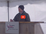 image of farmer standing a podium outside wearing a stocking cap and a red video play button